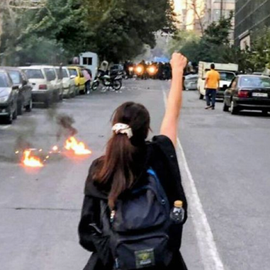 A person standing in the middle of a street with one arm raised, facing a line of vehicles and smoke from small fires on the road.